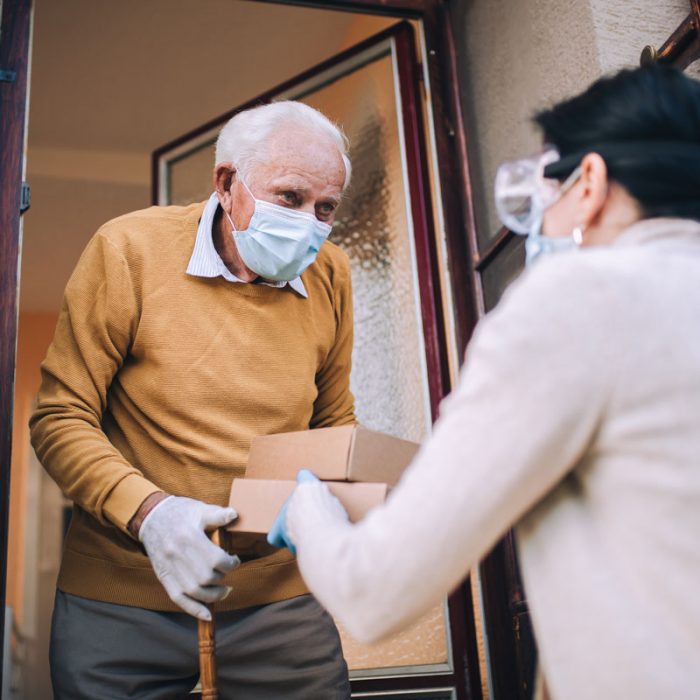 Young female volunteer in mask gives an elderly man a box of food as he is isolated during the covid-19 lockdown.